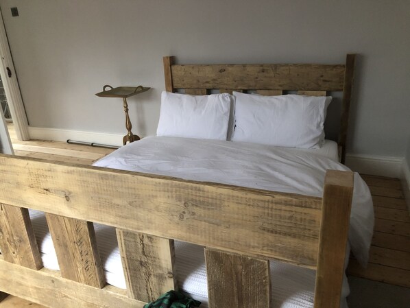 A wooden bed with white bedding in a room with a plain wall and a small table with a lamp. - Spacious & Tranquil Oasis in the Heart of Newquay; Spacious, Stylish, Coastal! (Newquay)