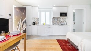 A modern hotel room with a wooden dining table set for two, a kitchen with white cabinets and a microwave, and a large bed with a red rug. - Lisbon Prime Aparts - Baixa (Lisboa)