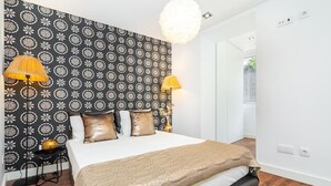 A bedroom with a patterned wallpaper, a bed with a beige blanket, two lamps, and a small table with a lamp.