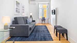 Un salon moderne avec un canapé gris, un tapis bleu, une table basse en verre et une lampe. On y trouve également un fauteuil et un téléviseur fixé au mur.