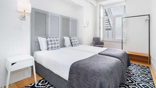 Une chambre moderne avec un grand lit, des tables de chevet, une chaise et une fenêtre donnant sur un immeuble.