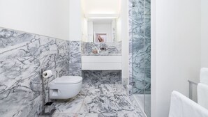 Une salle de bain aux murs et au sol recouverts de marbre, avec des toilettes blanches, une douche en verre et un lavabo blanc surmonté d’un miroir.