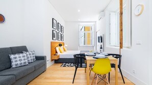 A modern apartment with a grey sofa, a dining table with chairs, a bed with a wooden headboard, and a large window. - Lisbon Prime Aparts - Baixa (Lisboa)