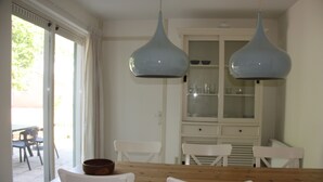 Un comedor con una mesa de madera y sillas blancas, dos lámparas colgantes azules y un aparador de vidrio con vajilla.