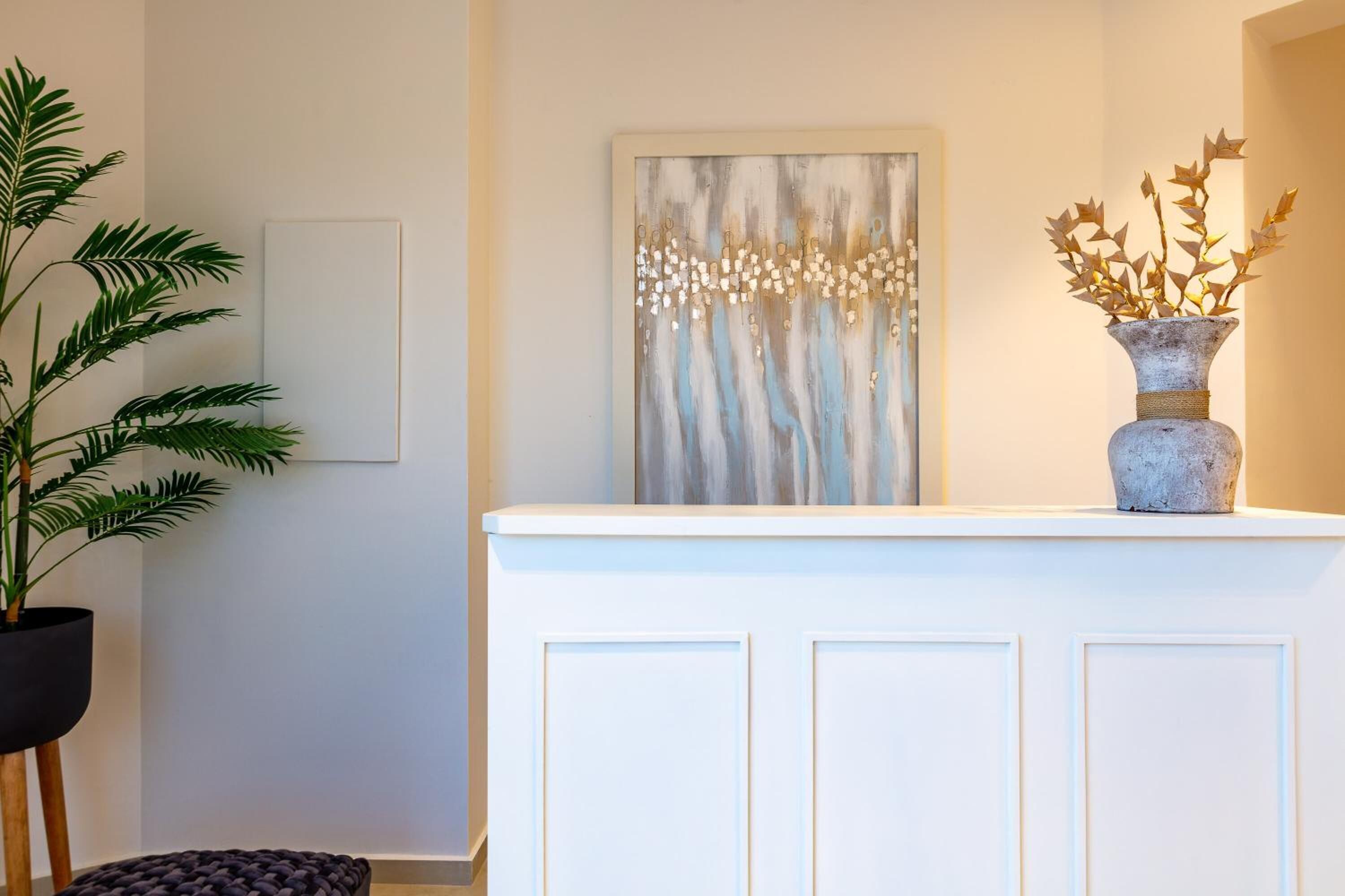 A reception area with a white reception desk, a potted plant, a framed artwork, and a vase with decorative branches.