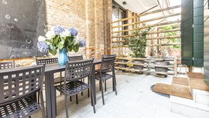 Patio esterno con muro in mattoni, tavolo e sedie, vaso blu con fiori e recinzione in legno.