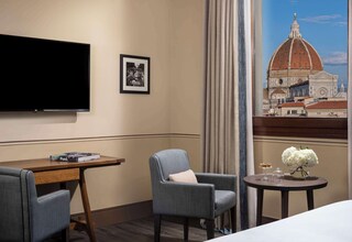 A hotel room with a flat-screen TV, a framed picture, a desk with a book, a chair, a small table with a vase of flowers, and a view of a historic building.