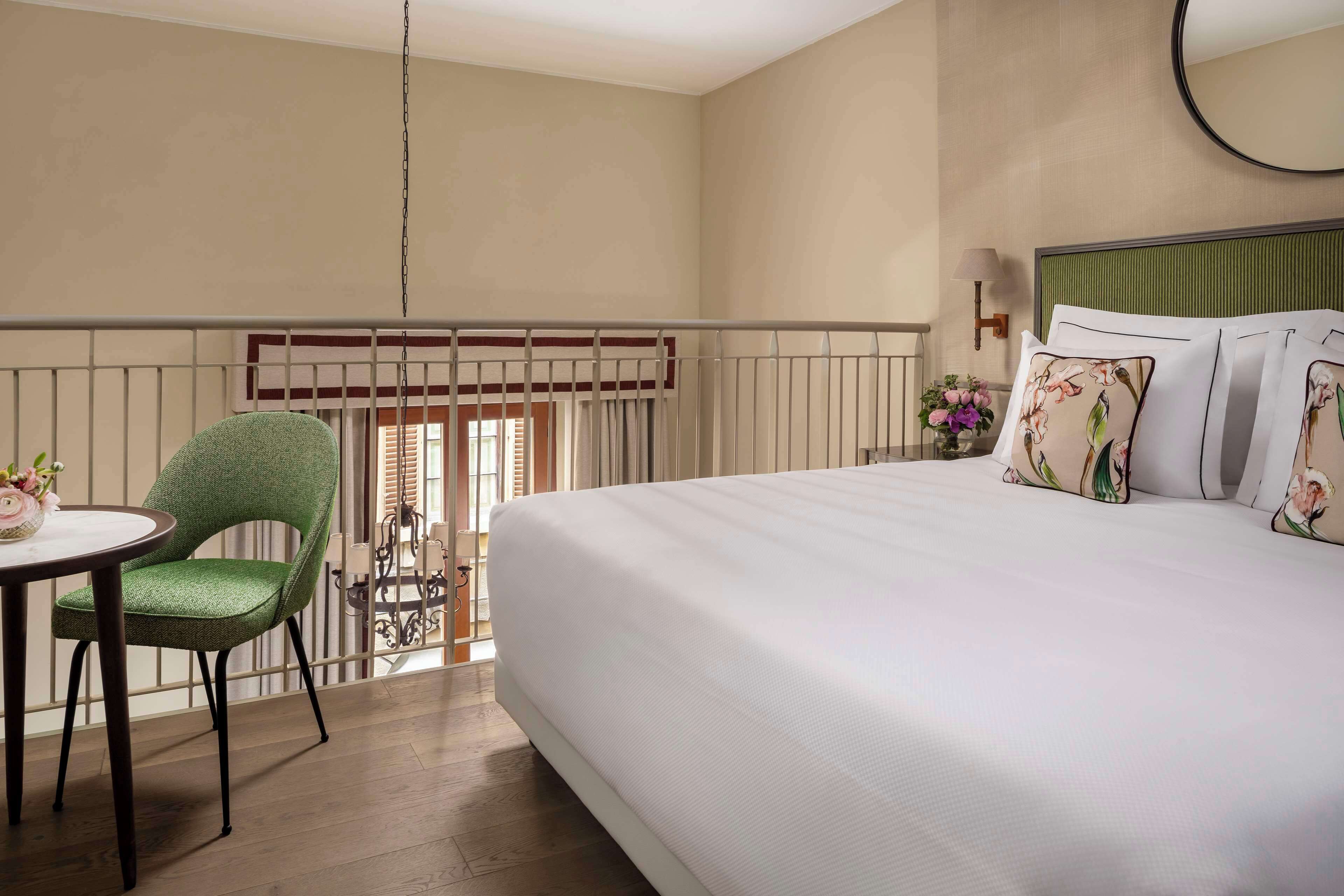 A hotel room with a bed, a green chair, a small table with a vase, and a window with blinds.