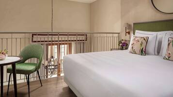 A hotel room with a bed, a green chair, a small table with a vase, and a window with blinds.