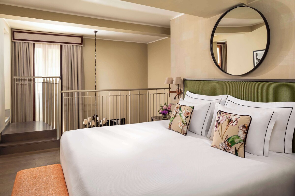 A hotel room with a large bed, a round mirror, a balcony with curtains, and a staircase.