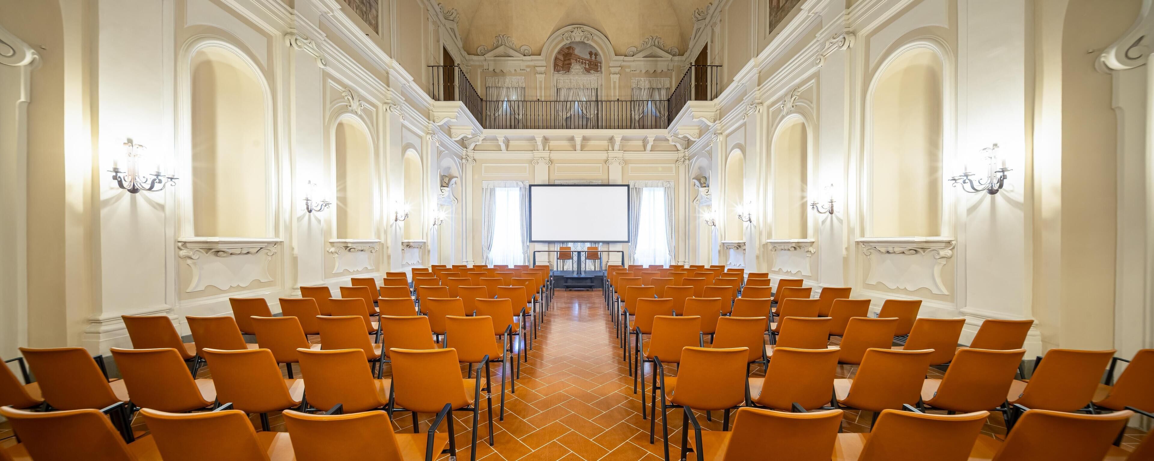 A conference room with rows of orange chairs, a large screen at the front, and ornate white walls with decorative moldings.