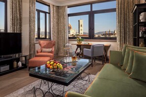 A modern living room with a glass coffee table, a sofa, a television, and a view of a cityscape through large windows. - Tivoli Palazzo Gaddi Firenze Hotel (Florence)