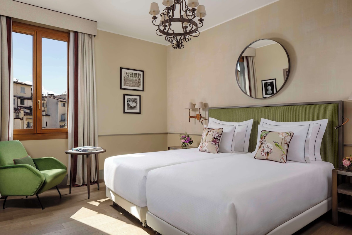A hotel room with a large bed, a green armchair, a small table, and a window with curtains.
