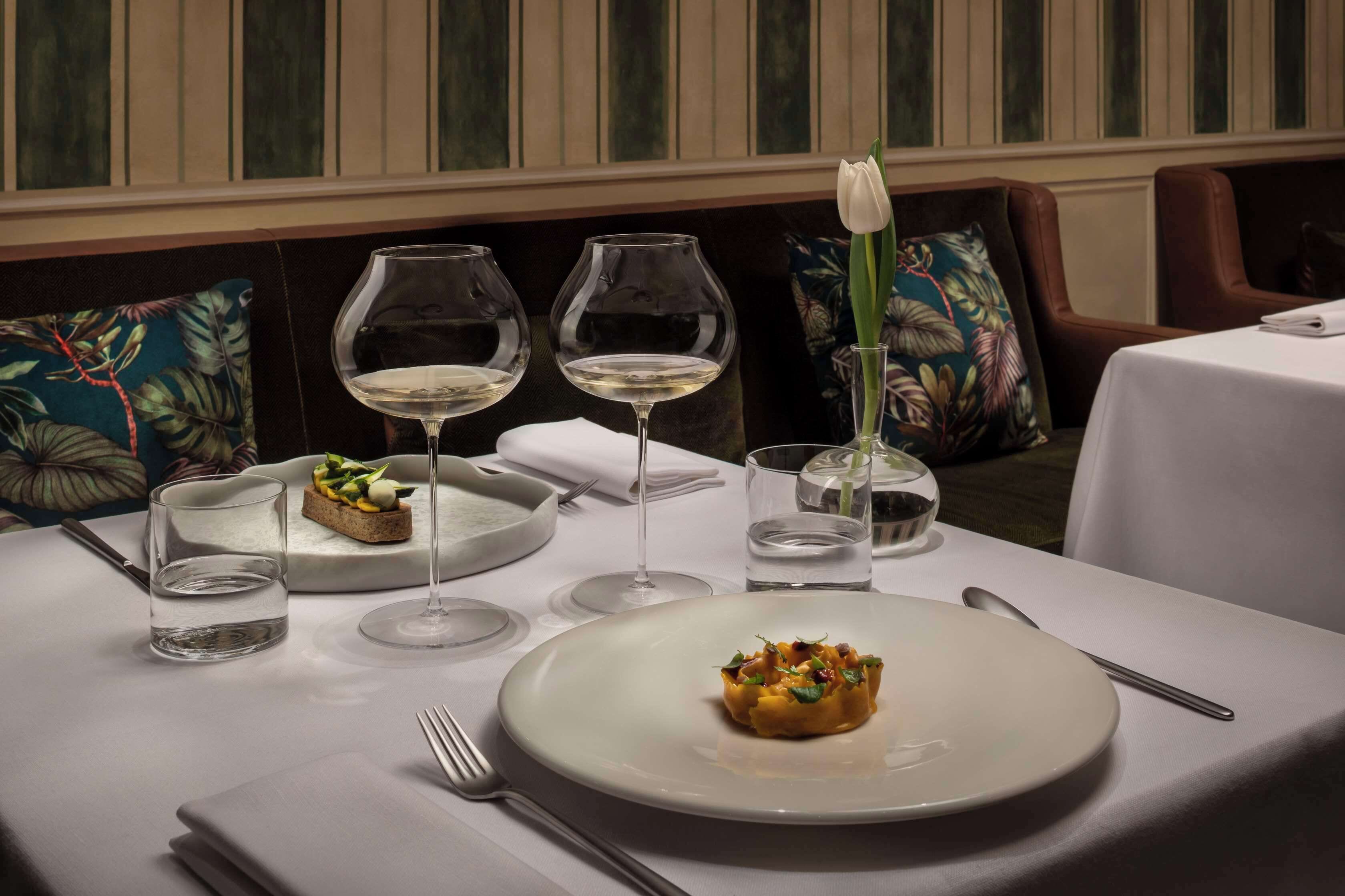 A dining table set with a plate of food, two wine glasses, a water glass, and a vase with a single flower.