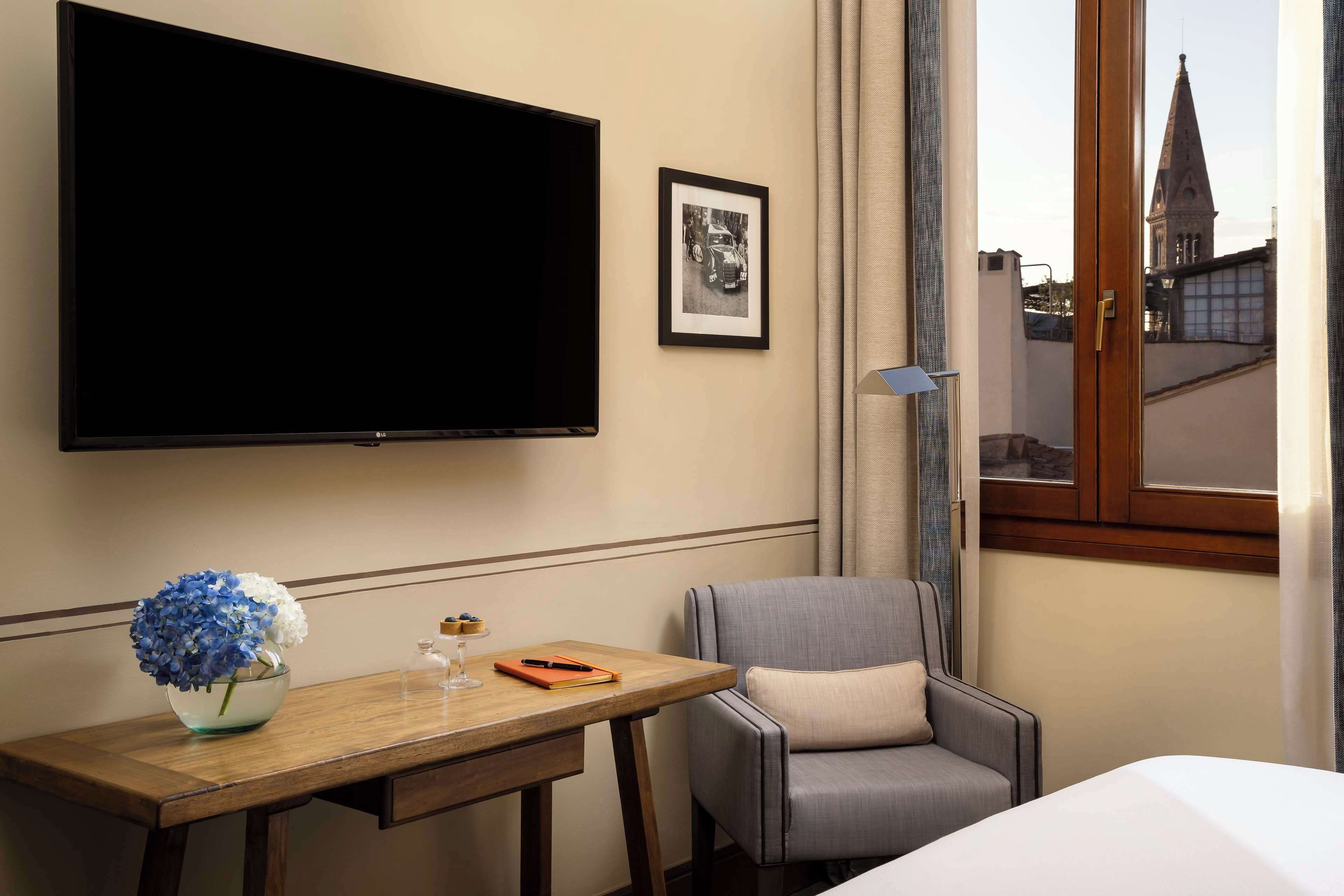 Una camera d'albergo con un letto, una scrivania con un vaso di fiori, una sedia, una TV e un quadro appeso al muro.