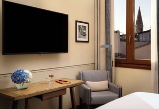 A hotel room with a bed, a desk with a vase of flowers, a chair, a TV, and a framed picture on the wall.