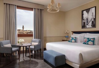 A hotel room with a large bed, two armchairs, a small table with a vase of flowers, and a view of a tower through the window.