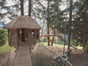 A wooden treehouse with a rope bridge and a slide, surrounded by tall trees. - Hieserhof (Neustift Im Stubaital)