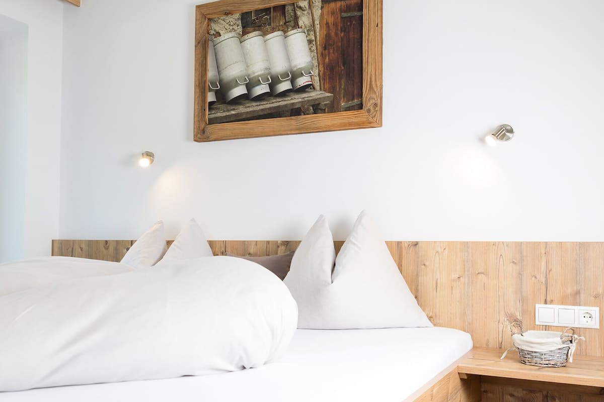 A bedroom with a wooden headboard, white bedding, a small bedside table with a basket, and a wall-mounted light fixture.