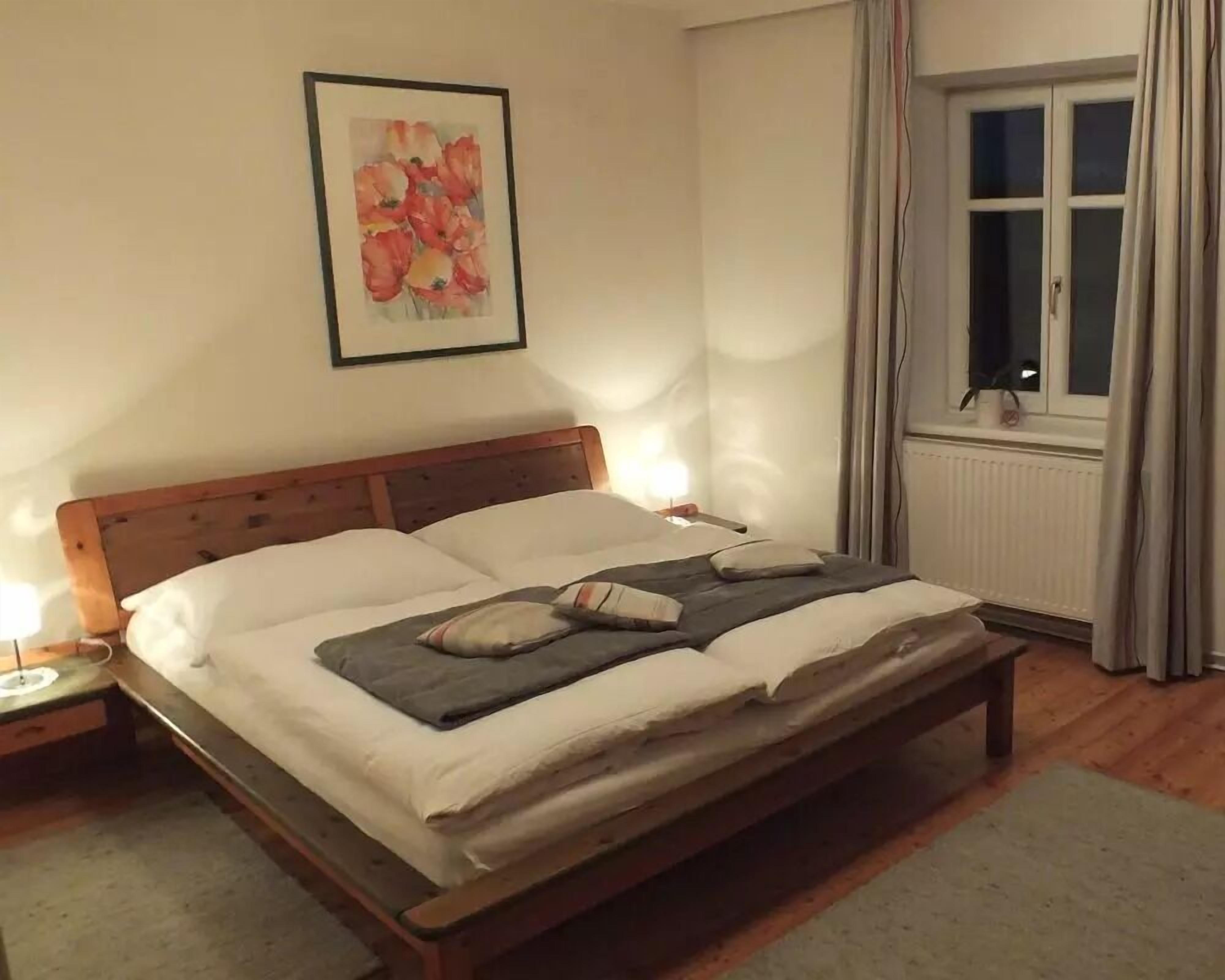 A neatly made bed with pillows and a blanket, a bedside table with a lamp, a framed floral artwork on the wall, and a window with curtains.