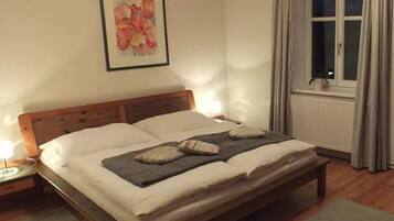 A neatly made bed with pillows and a blanket, a bedside table with a lamp, a framed floral artwork on the wall, and a window with curtains.