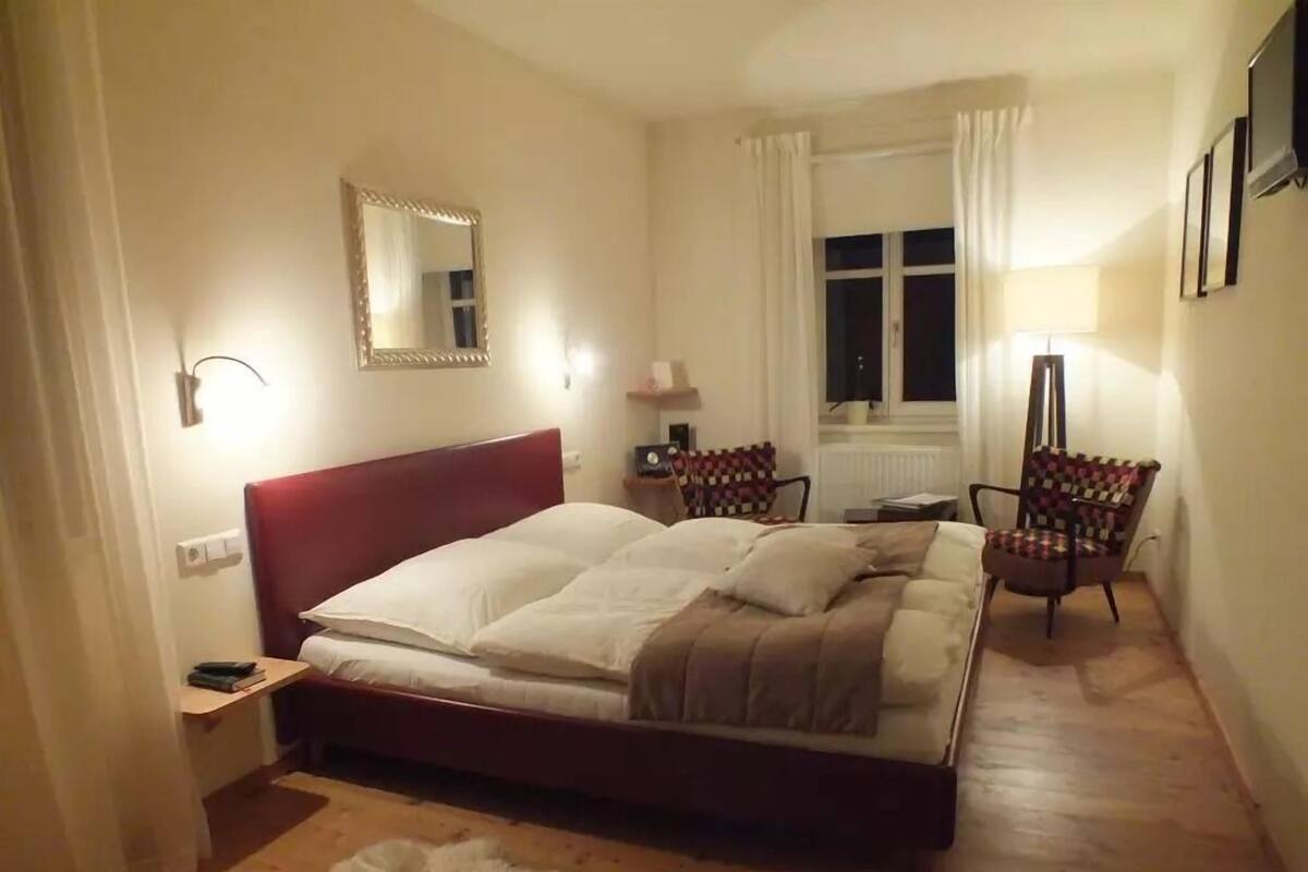 Ett sovrum med en stor säng, ett sängbord, en stol, en spegel och ett fönster med gardiner.
