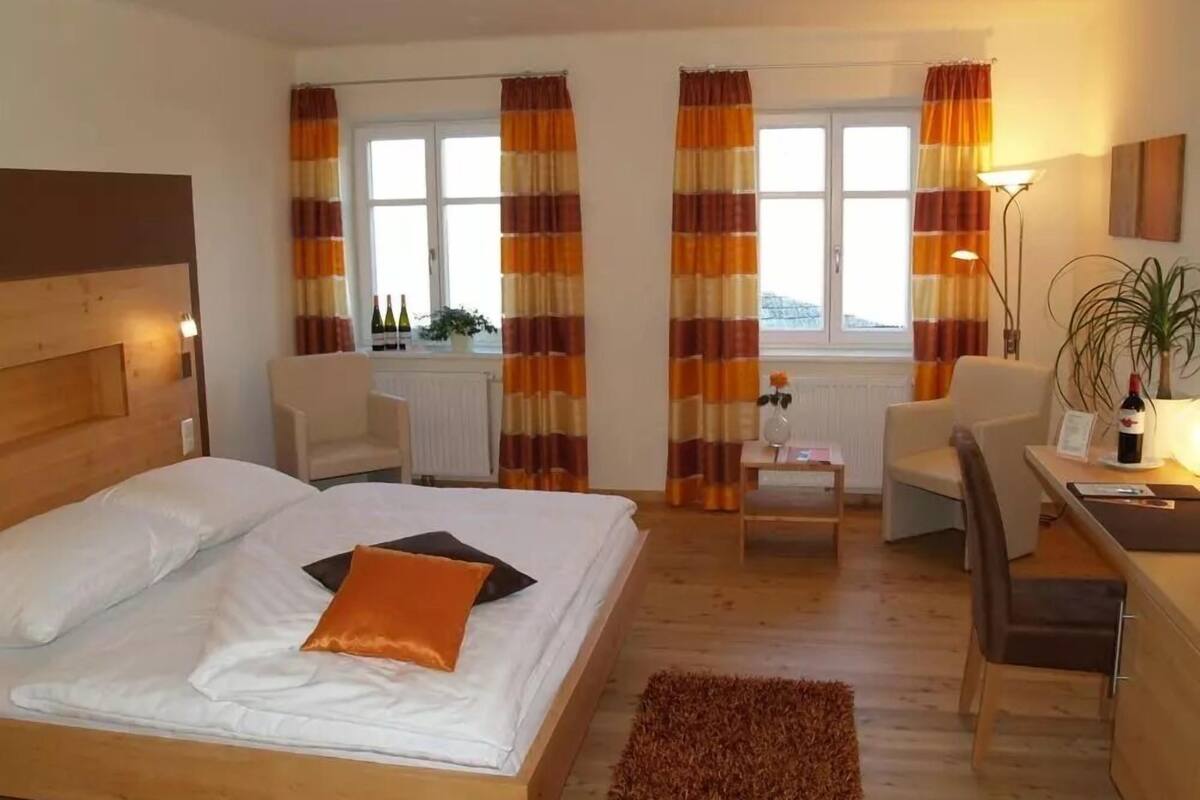 Ett hotellrum med en säng, två stolar, ett litet bord, en lampa och ett fönster med gardiner.