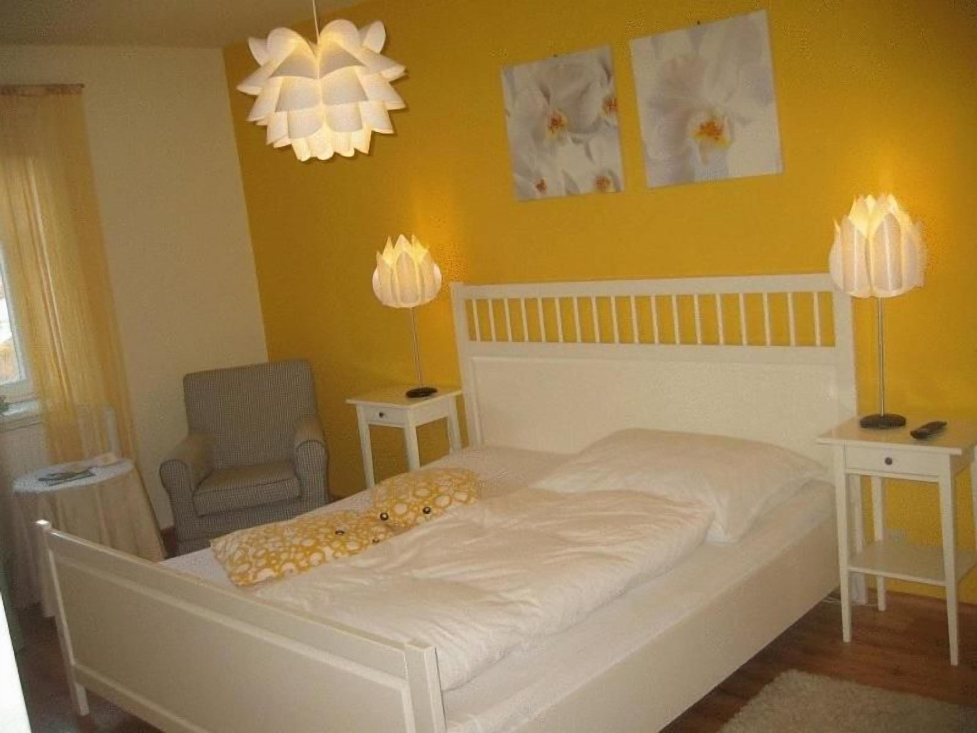 Una cama con ropa de cama blanca, dos lámparas de noche y una silla en una habitación con paredes amarillas.