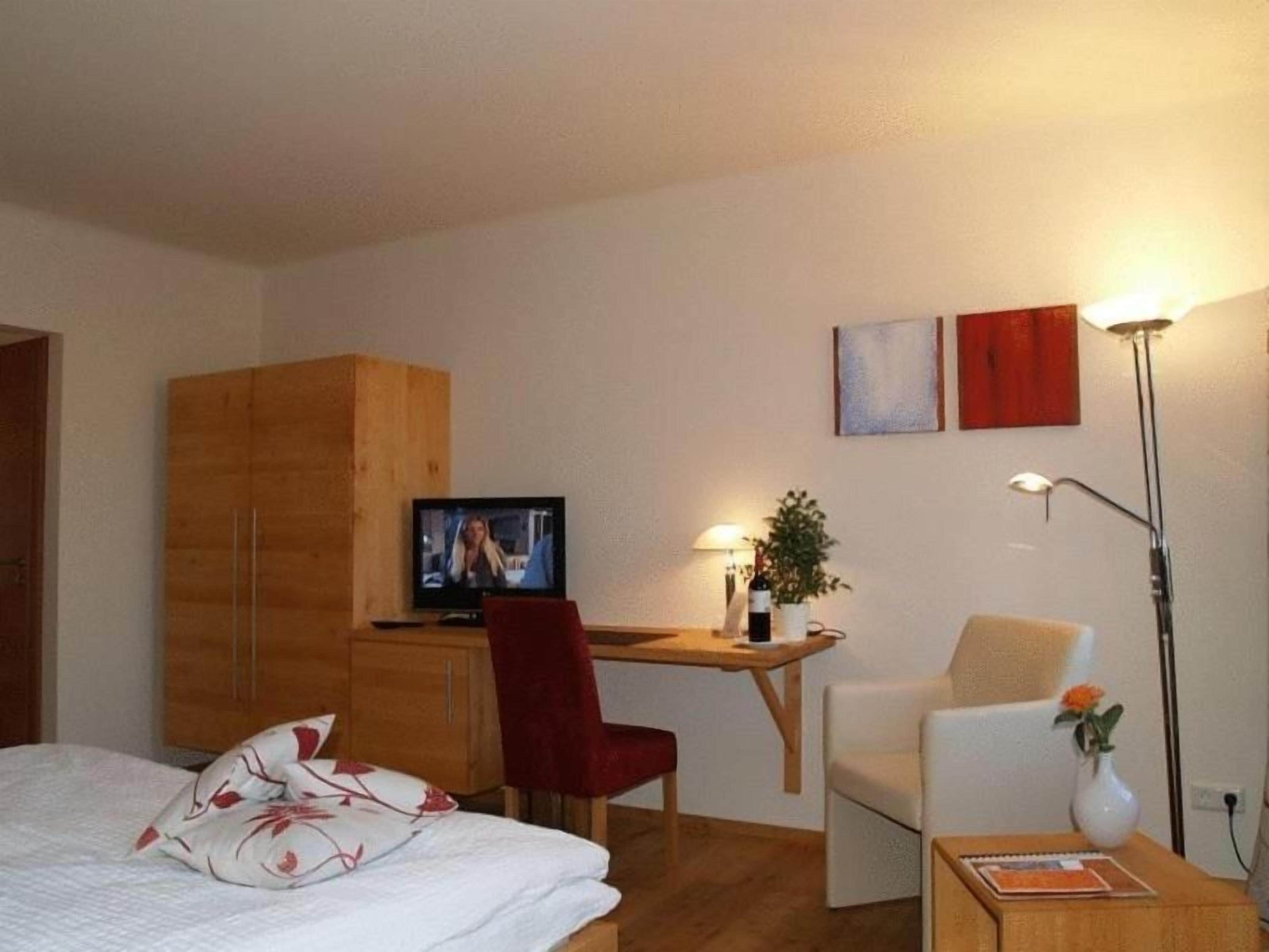 Una habitación de hotel con una cama, un escritorio con televisor, una silla, una mesita con un florero y una lámpara.