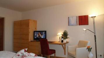 Una habitación de hotel con una cama, un escritorio con televisor, una silla, una mesita con un florero y una lámpara.