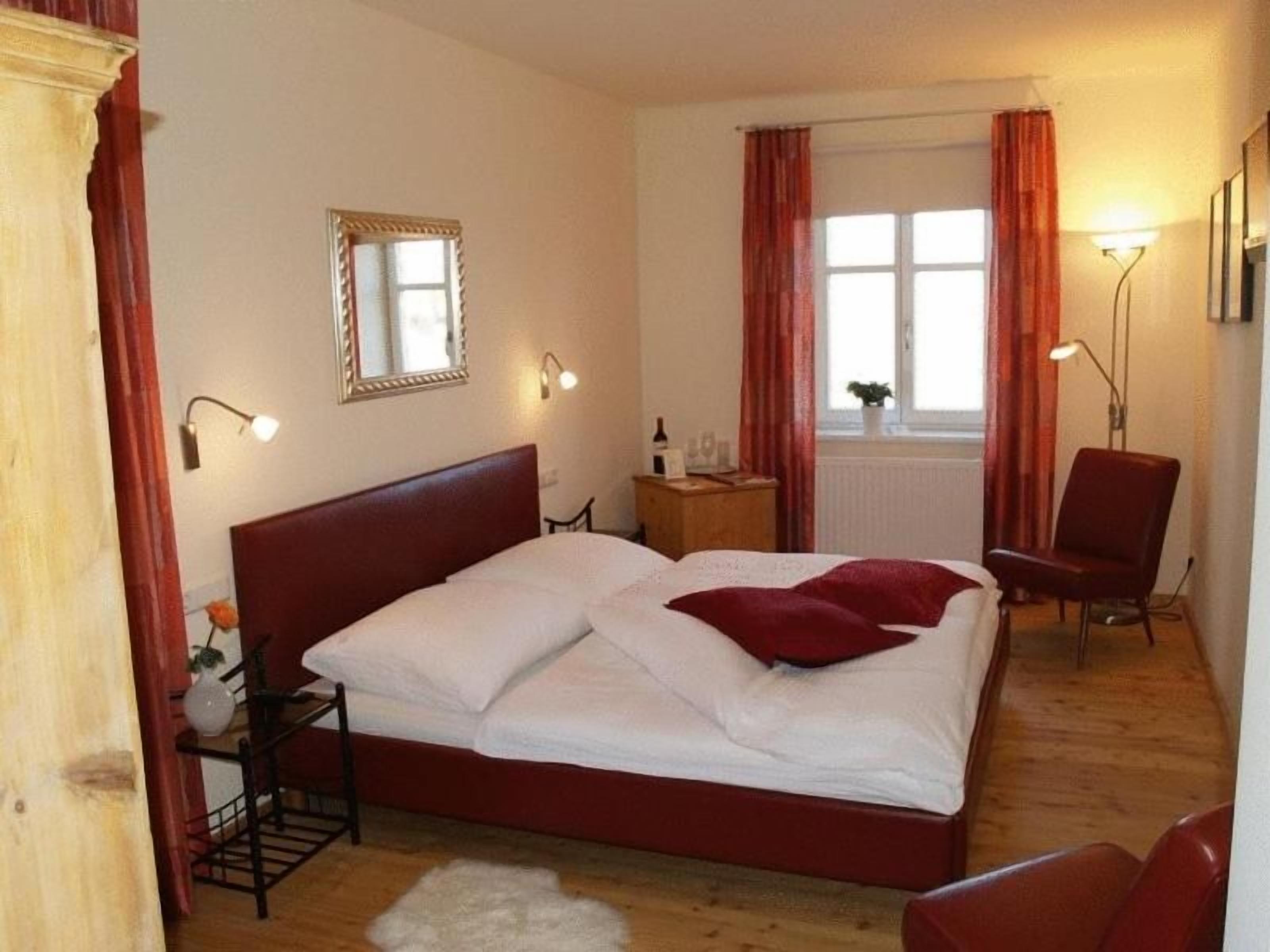 Un dormitorio con una cama grande, mesitas de noche, una silla, una ventana con cortinas y lámparas de pared.