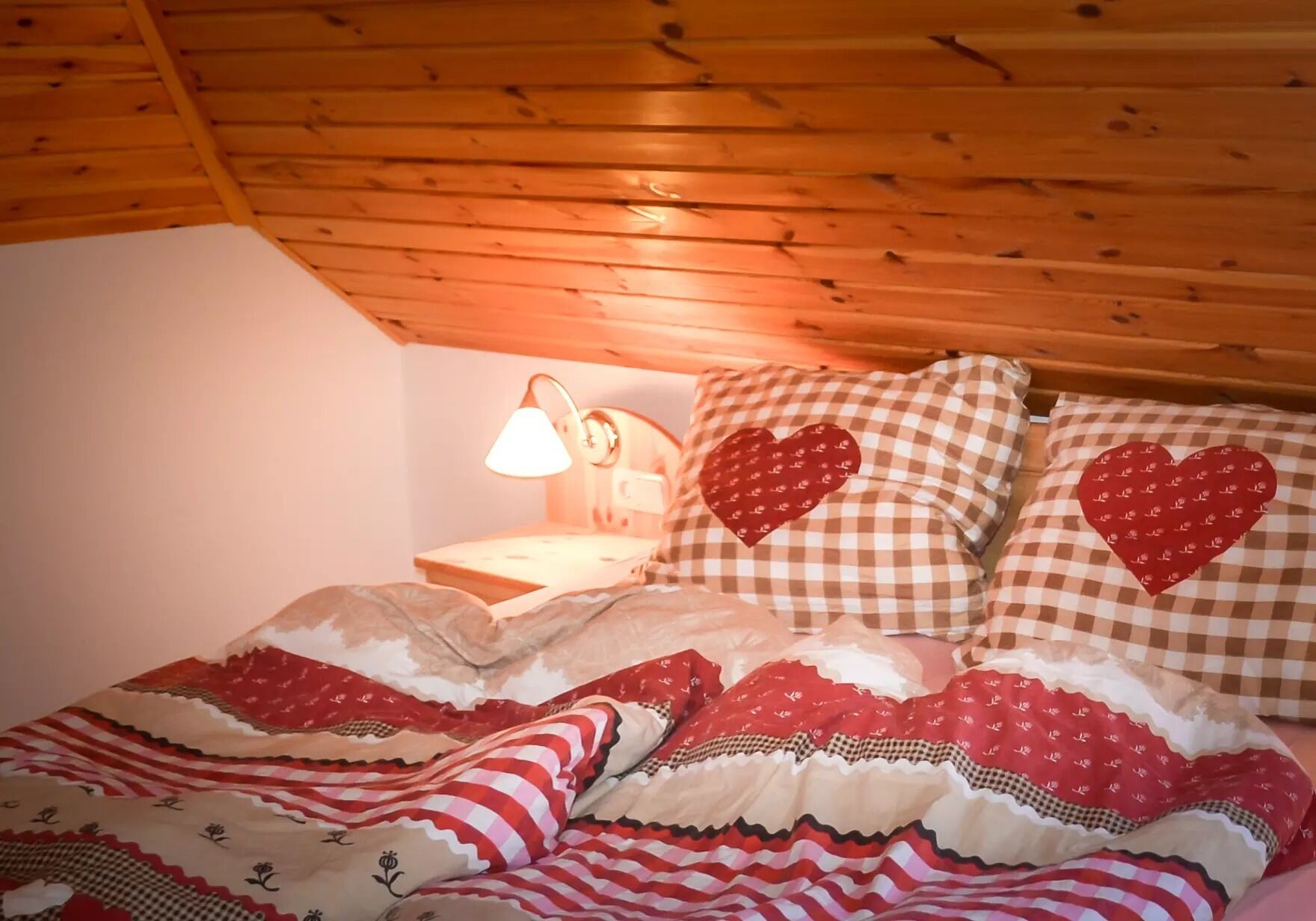 Una habitación acogedora con techo de madera, cama con almohadas a cuadros y un diseño de corazón rojo, además de una lámpara de noche.