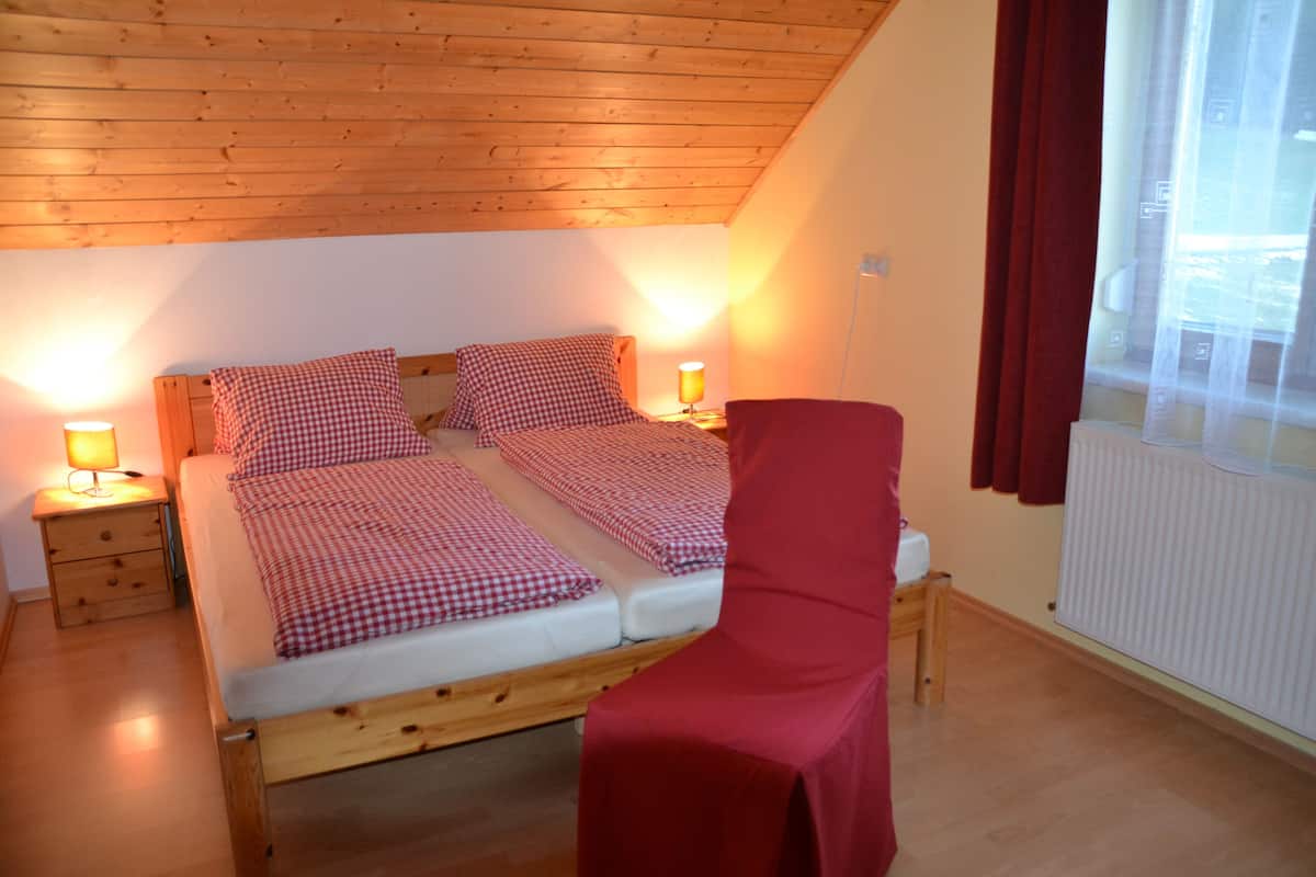 A bedroom with a wooden ceiling, two beds with checkered bedding, bedside lamps, a red chair, and a window with curtains.