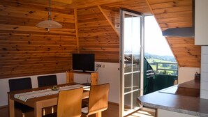 Ein Zimmer mit Holzwänden, einem Esstisch, Stühlen, einem Fernseher und einer Glasschiebetür, die auf einen Balkon mit Blick auf die Landschaft führt.