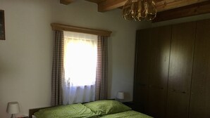 Un dormitorio con cama, armario, ventana con cortinas y mesita de noche con lámpara.