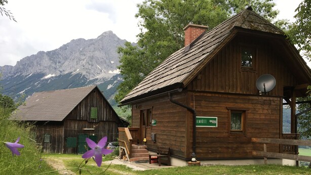 Una cabaña de madera rústica con antena parabólica, un letrero que indica 'Almhütte' y un camino de tierra que la lleva.