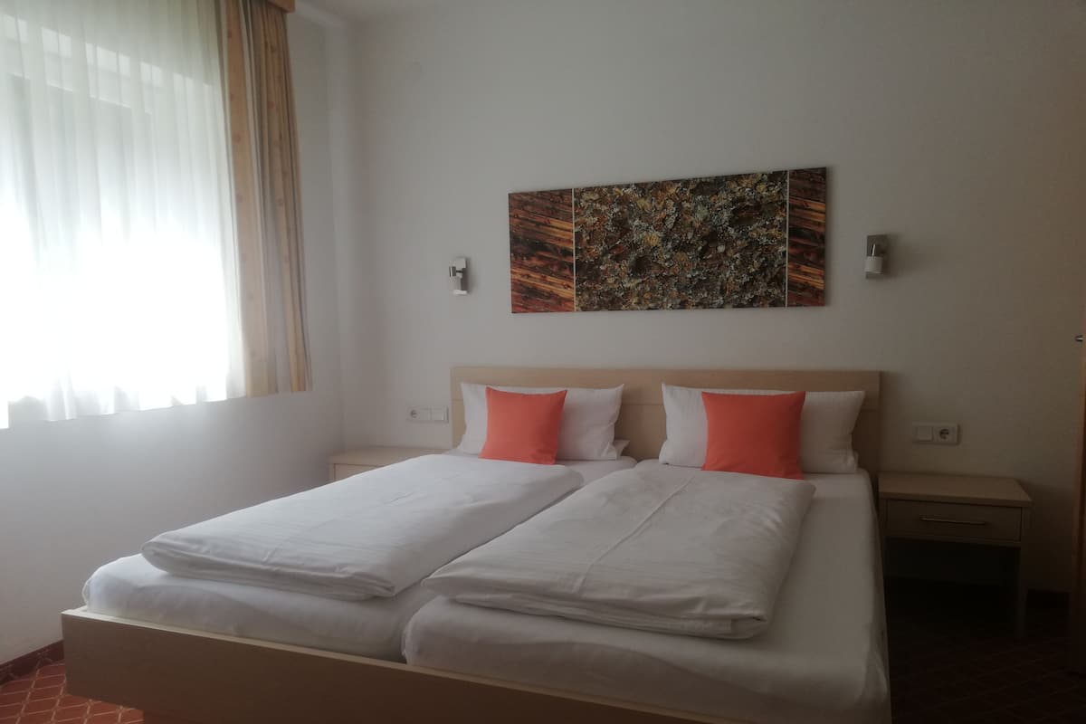 Cama doble con sábanas blancas y dos almohadas naranjas, mesita de noche con lámpara y un cuadro en la pared.