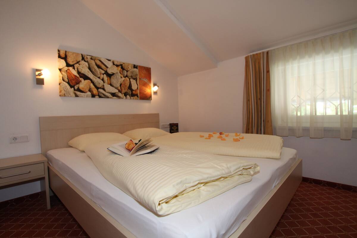 Una cama bien hecha con un libro, una obra de arte en la pared y una ventana con cortinas.