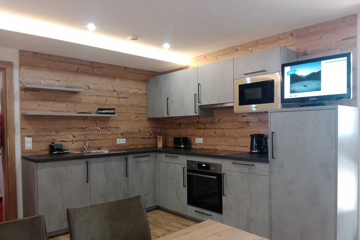 Una cocina moderna con armarios grises, horno empotrado, microondas y un televisor encima de la nevera.