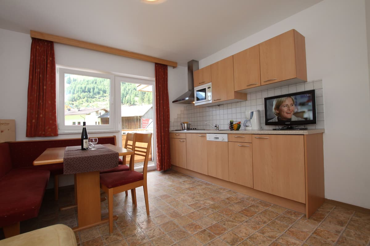 Cocina compacta con armarios de madera, un microondas y un televisor. Hay una zona de comedor con una mesa y sillas, y una ventana con cortinas.
