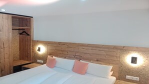 Un dormitorio con cabecera de madera, dos lámparas de noche, cama con ropa de cama blanca y almohadas rosas, y un armario de madera con perchero.