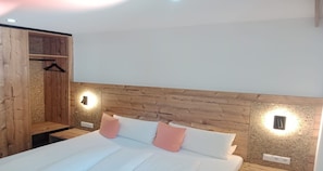 A bedroom with a wooden headboard, two bedside lamps, a bed with white bedding and pink pillows, and a wooden wardrobe with a hanging rack.