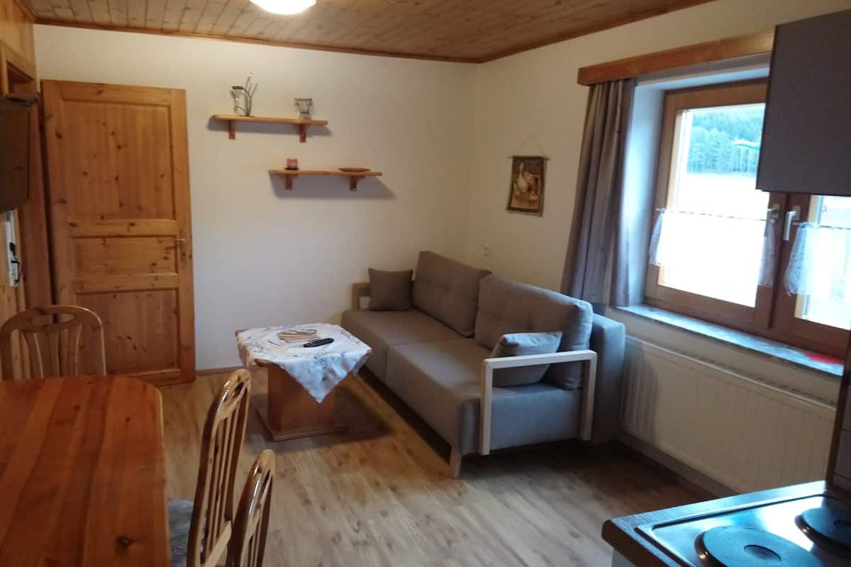 A cozy living room with a wooden dining table, a sofa, and a small table with a white tablecloth. There are wooden shelves on the wall and a window with curtains.