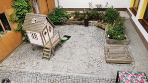 Un patio con una casita de juegos de madera, una mesa pequeña con sillas y plantas en macetas.