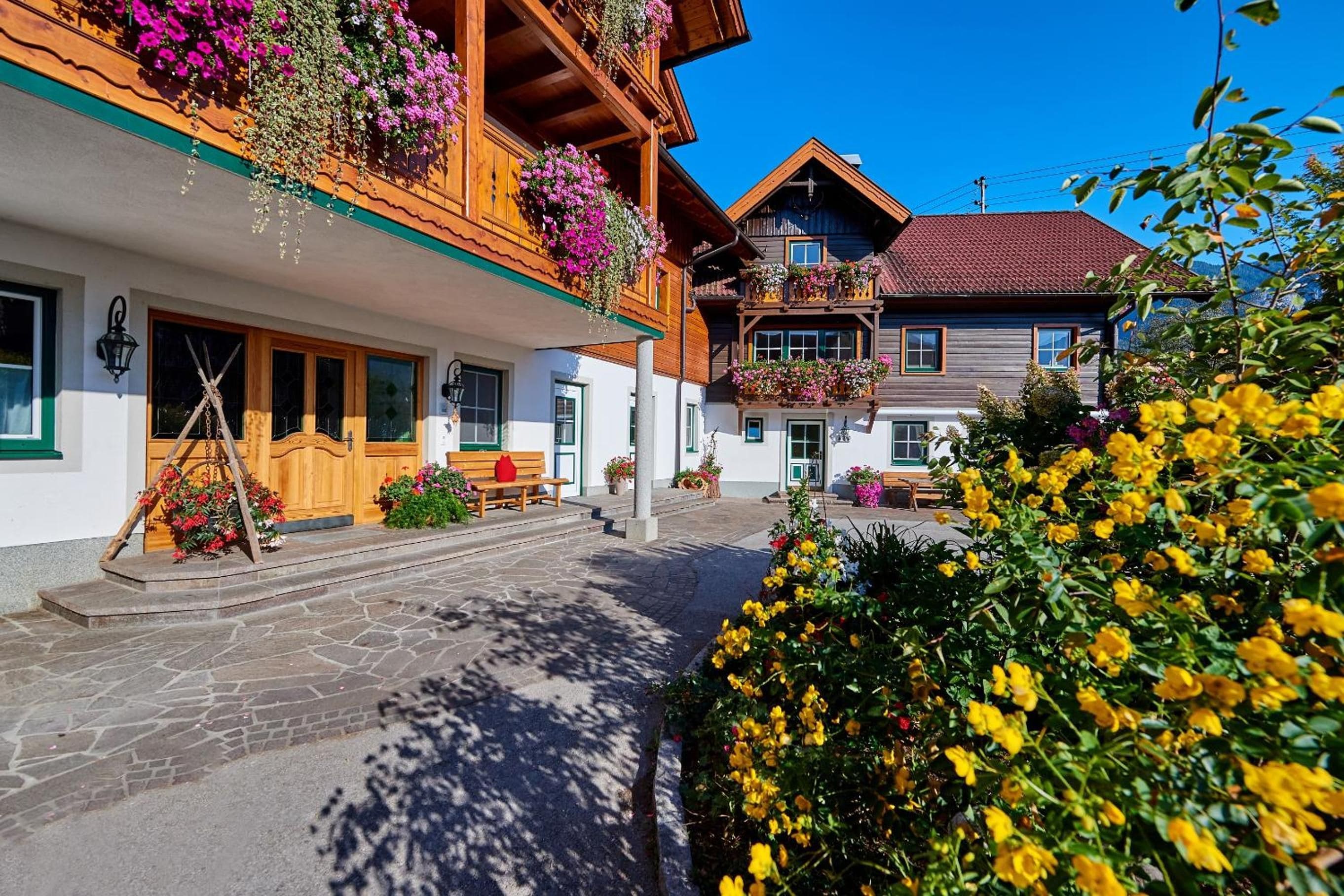 Ein traditionelles Holzgebäude mit Balkonen, die mit Blumen geschmückt sind, eine gepflasterte Gasse und ein klarer, blauer Himmel.