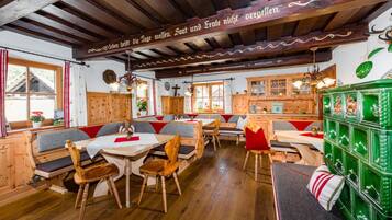 A traditional inn with wooden beams, a central seating area with benches and tables, and a green cabinet with glassware.