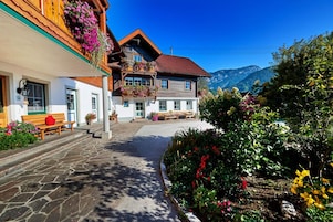 A traditional chalet with a stone-paved entrance, wooden balconies with flower boxes, and a mountain backdrop.
