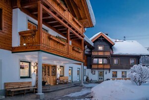 A traditional wooden chalet with a balcony, a snow-covered roof, and a bench outside.
