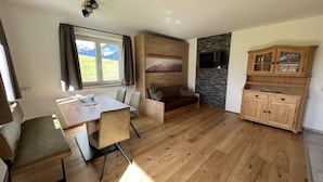 A cozy room with a wooden dining table, chairs, a sofa, and a TV mounted on a stone wall. - Studlerhof (Oberperfuss)
