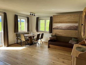 A room with a wooden dining table, four chairs, a wooden sofa, and a large window with a view of greenery.